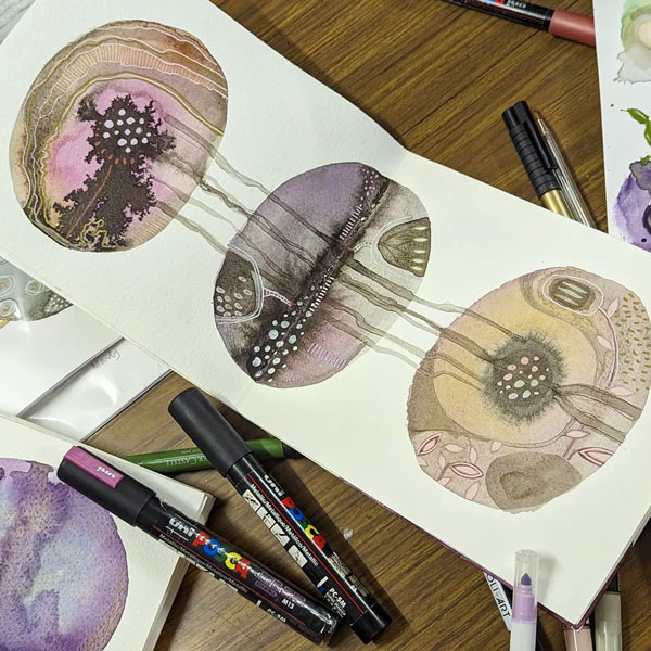 a modern watercolour design in a sketchbook surrounded by markmaking tools and art supplies